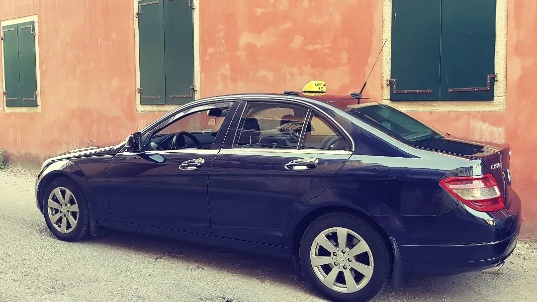 The dark blue Mercedes taxi in front of an old Corfiot house with green shutters